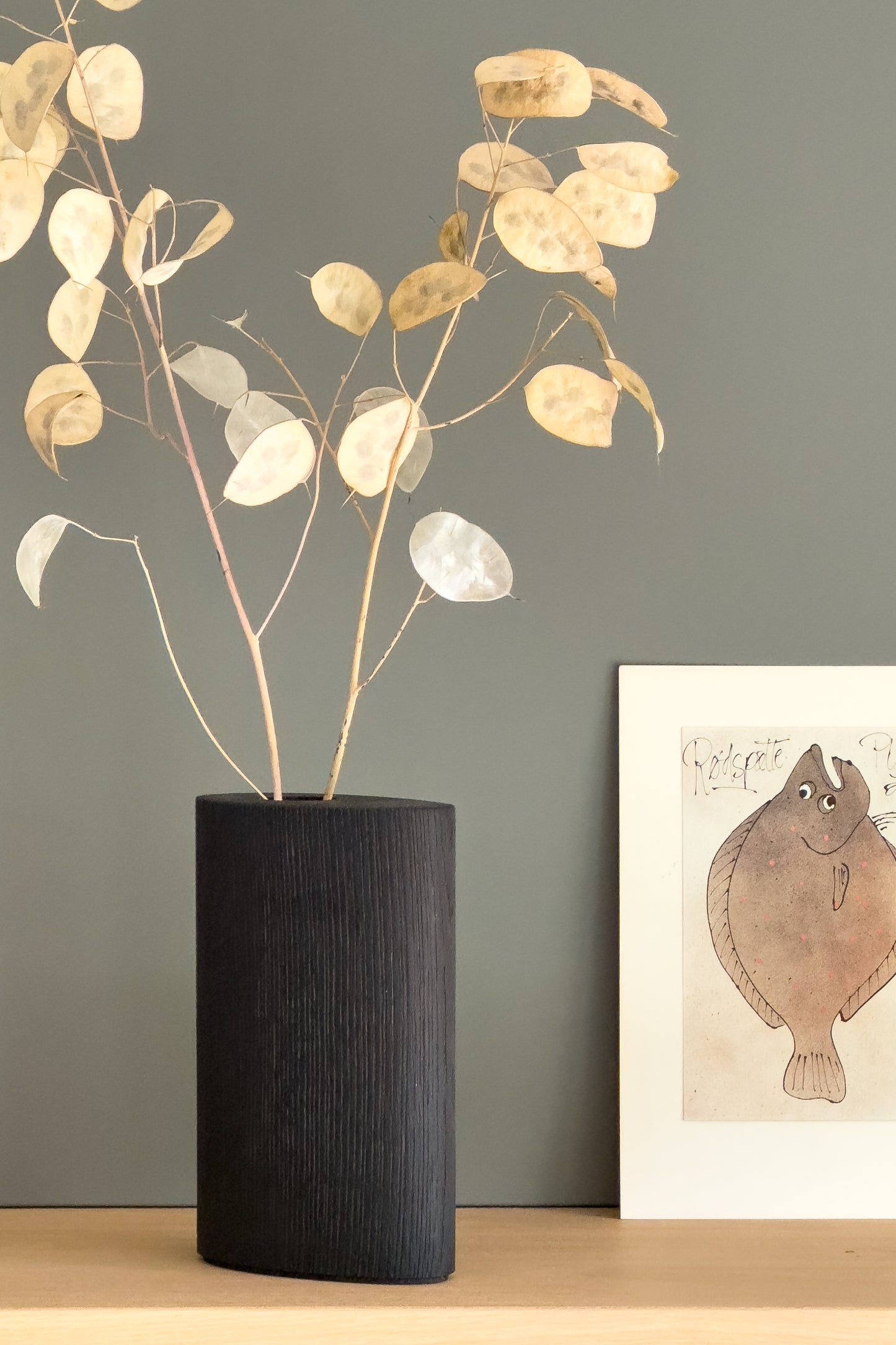 Unsere moderne, handgefertigte Holzvase GROW! Die Vase aus dunkler Eiche im Farbton „Bold“ steht hier auf einem Regalboden aus heller Eiche. In Kombination mit gedeckten Erdtönen und der Trockenpflanze Silberblatt wirkt sie minimalistisch und zeitlos. Stilvolle und hochwertige Dekoration für dein modernes Wohnambiente.
