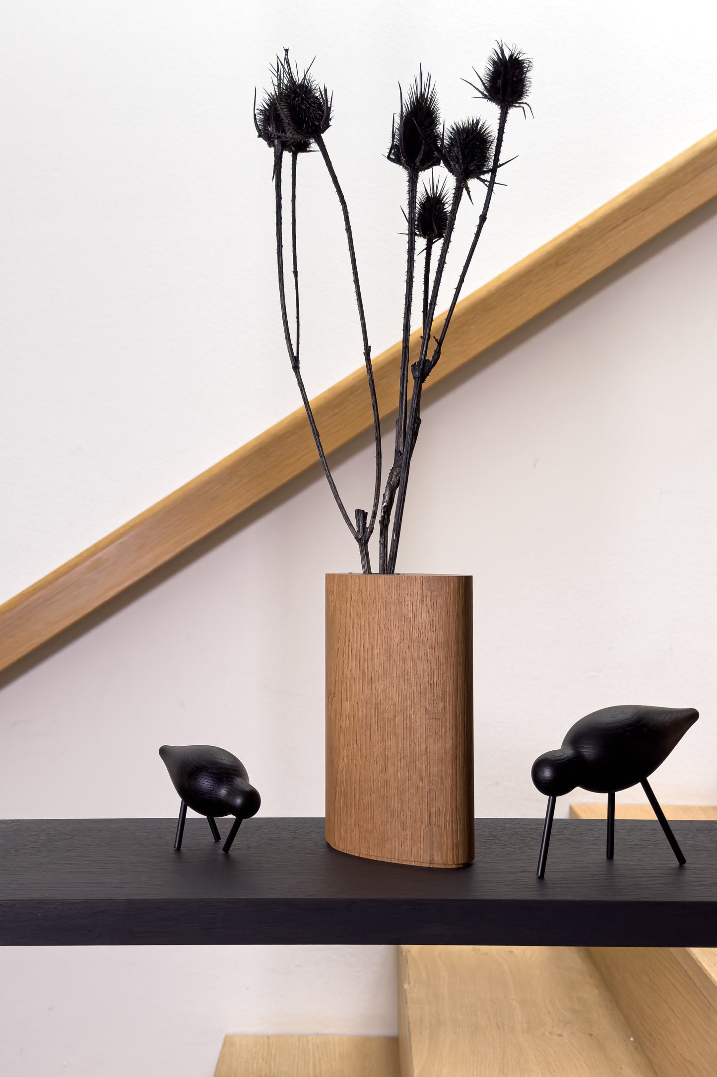 Die Vase in naturfarbener Eiche steht auf einem Regalbrett neben Vogelfiguren von Normann Copenhagen. Sie bildet einen Hell-Dunkel-Kontrast zum dunklen Holzton der Figuren und des Regalbodens sowie zur schwarz eingefärbten Trocken-Distel. Im modernen Treppenhaus-Kontext wirkt sie stilvoll und zeitgemäß