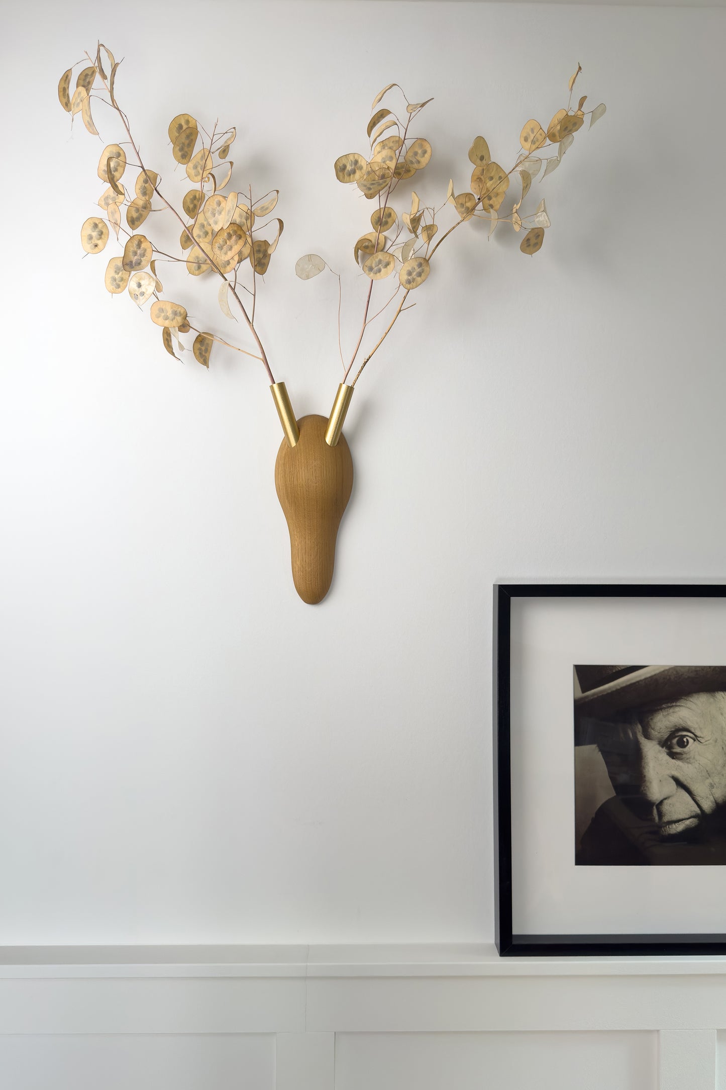 Das Bild zeigt eine Wandverkleidung, auf der ein Bilderrahmen mit einem Portrait Picassos steht. Unsere Wanddekoration zeigt RESPECT! Eine tierfreie Geweihinterpretation, in deren goldenen Vasenrörchen getrocknetes Silberblatt steckt .