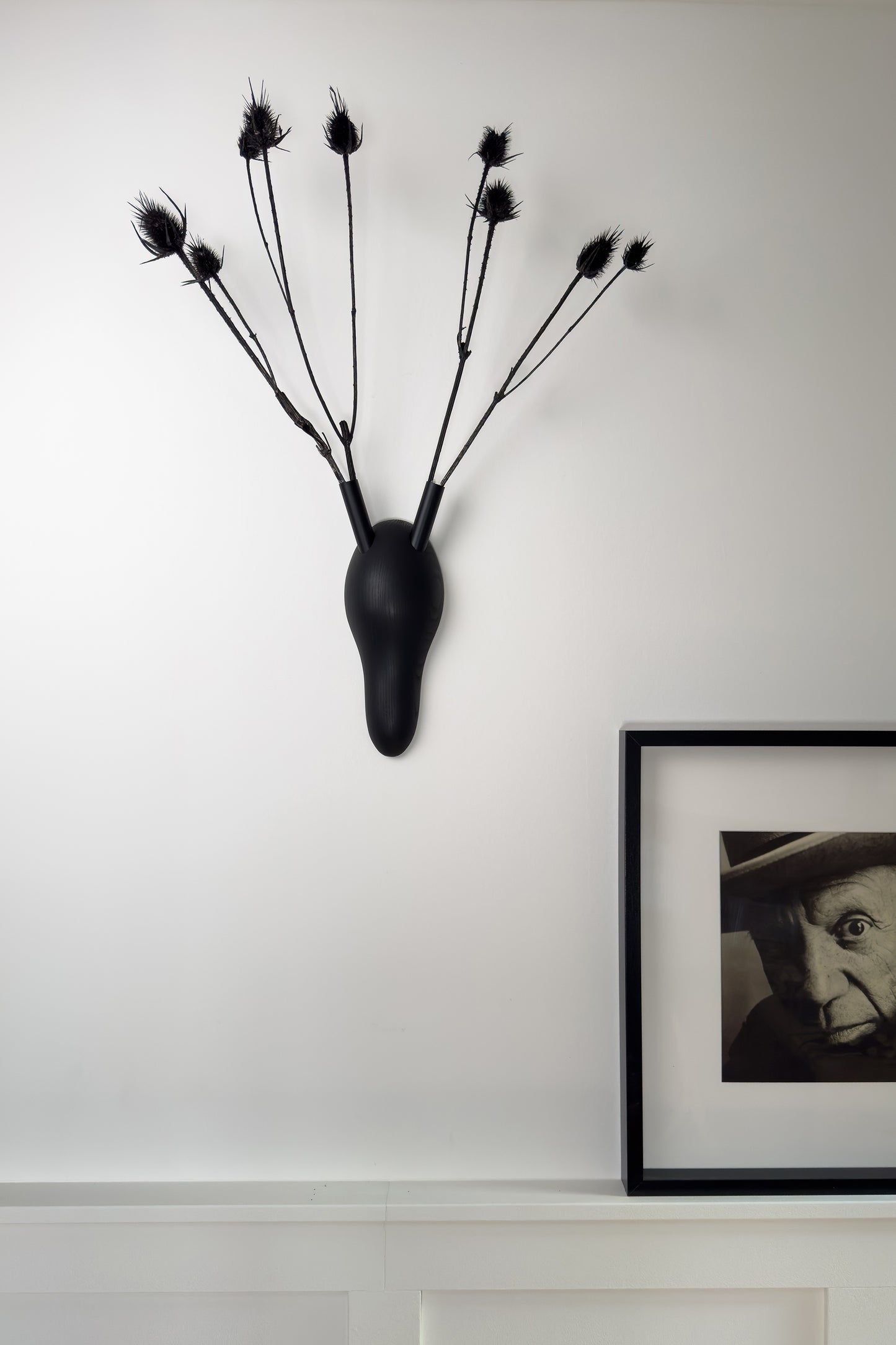 Unser Wandschmuck aus gedrechseltem Massivholz ist ein tierfreies Geweih. In den "Hörnern" finden frische Blumen oder Trockenpflanzen Platz. Unser uni schwarzes Modell trägt im Bild schwarze Disteln. Ein Portrait von Pablo Picasso steht auf einer Wandverkleidung und komplettiert das Bild.