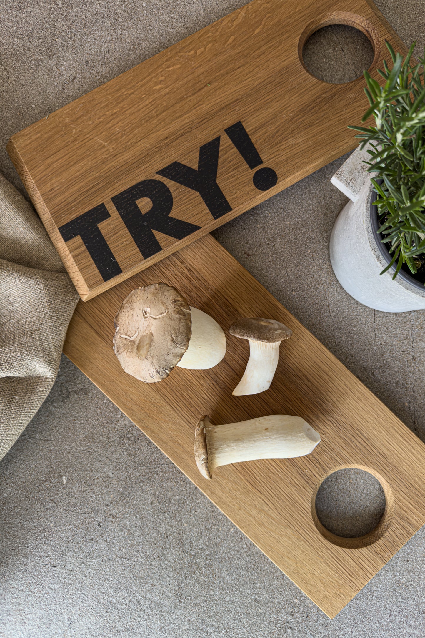 TRY! Unser Schneidebrett ist dein Küchenhelfer für die hygge Küche. Auf dem Foto ist das Servierbrett von der Ober- und Unterseite abgebildet. Auf der Unteseite steht der motivierende Schriftzug TRY! Auf dem anderen Brett liegen drei Kräuterseitlinge und Rosmarin in einem Kräutertopf vervollständigen das Bild.