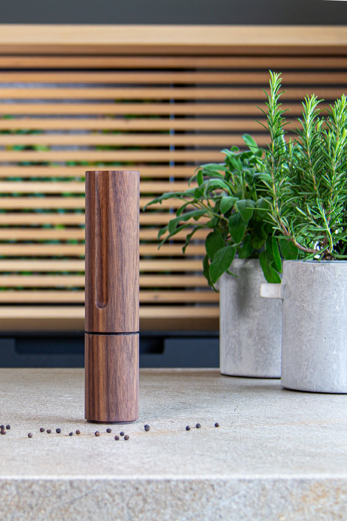 Handgefertigte Pfeffermühle aus Nussbaum in modernem Design und hochwertiger Technik. Umgeben von zwei Kräutertöpfen und einer Lamellenoptik im Hintergrund. Stilvoll für deine exklusive Küche und Kochkunst. Auch ein super Weihnachtsgeschenk!