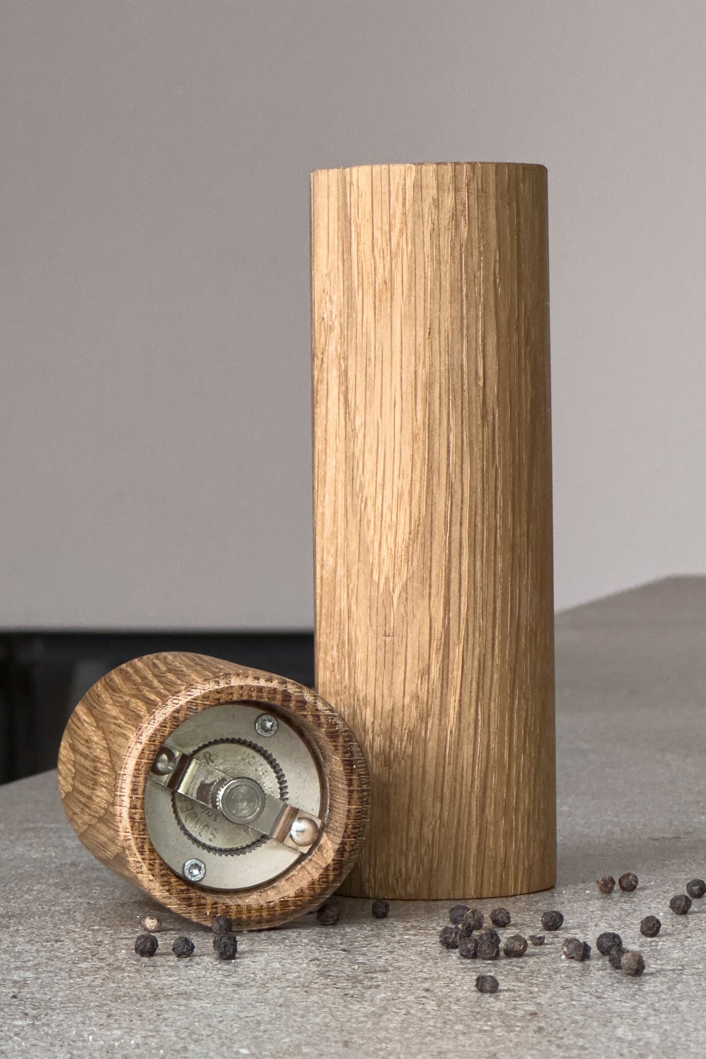 Design-Pfeffermühle aus Holz mit Blick auf das Mahlwerk, dekoriert mit Pfefferkörnern auf einer Steinarbeitsplatte. Ansicht des gehärteten Stahlmahlwerks für feinste Einstellung des Mahlgrades. Profiequipment für deine Küche!