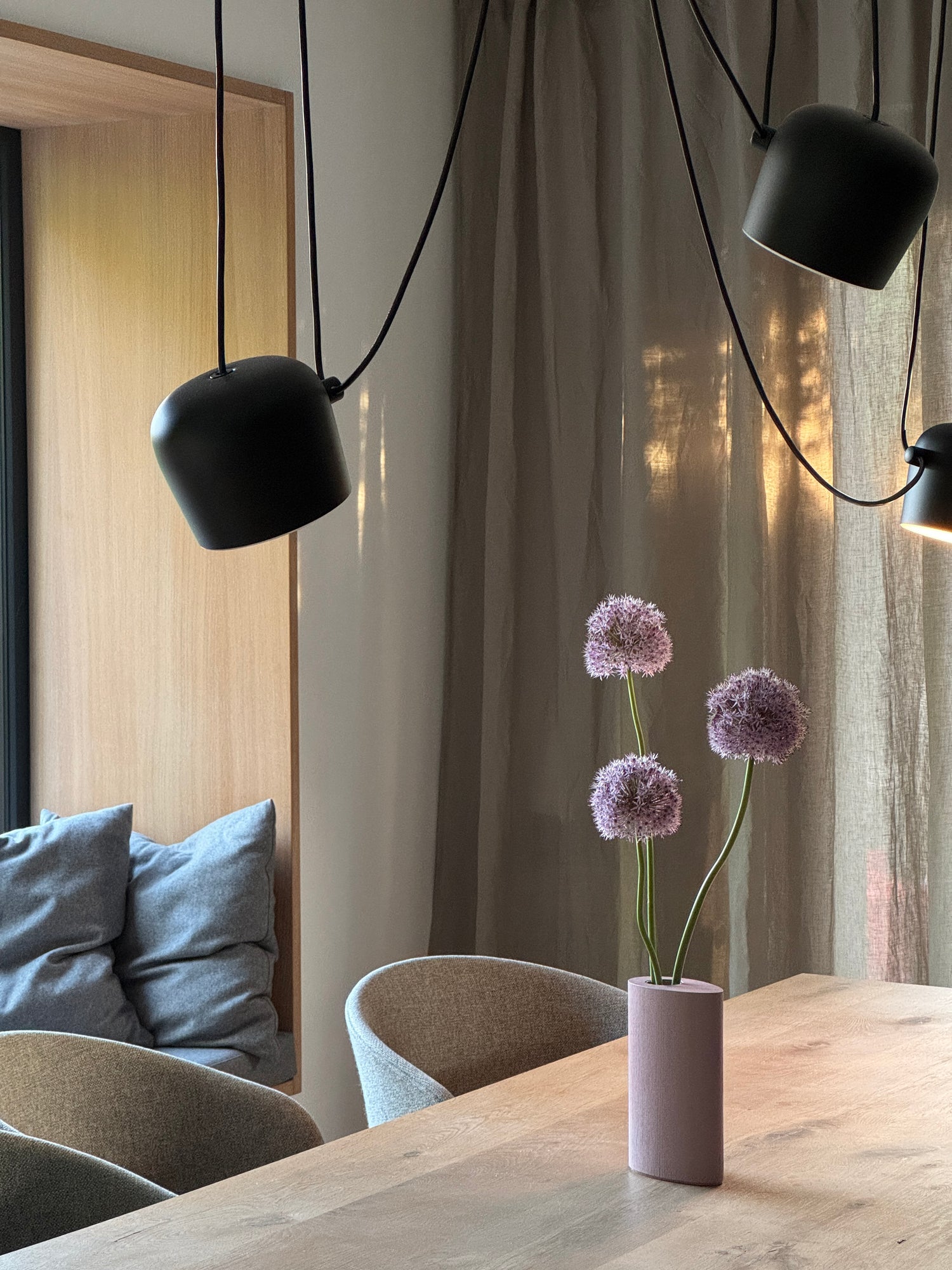 Holzvase GROW! mit Allium auf einem Holztisch in warmer Slow Living Atmosphäre in einer Nordic Design Einrichtung. Sonnenlicht  scheint durch ein Sitzfenster und durchdringt diffus einen Vorhang. Über dem Tisch hängt die Pendelleuchte Aim von Flos.