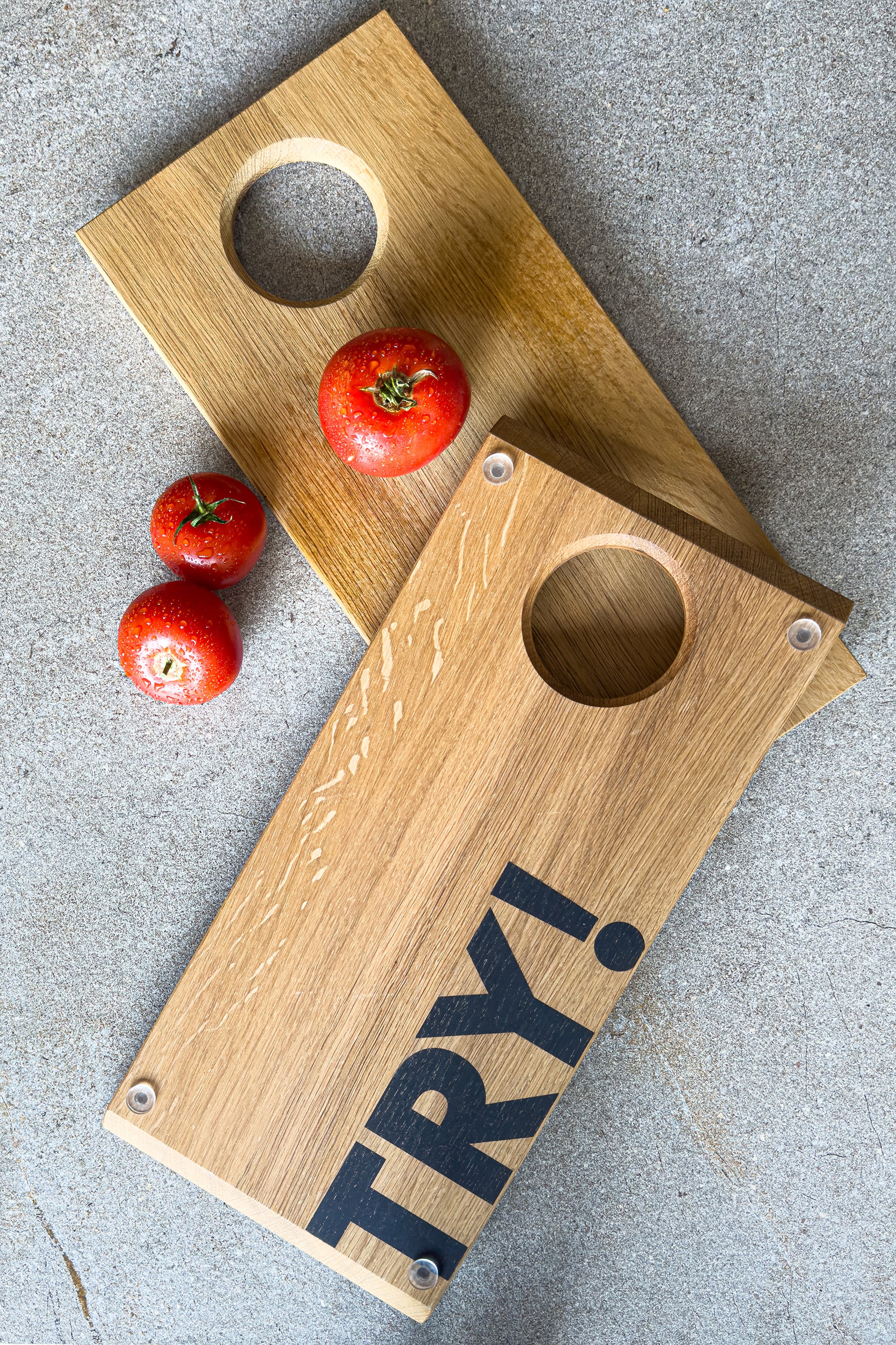 TRY! Holzschneidebrett mit Motivationstext auf der Unterseite! Dargestellt sind Ober- und Unterseite des nachhaltigen Küchenaccessoires und drei frischen Tomaten auf grauem Steinuntergrund. Hochwertiges Eichenbrett mit modernem Design und praktischer runder Aussparung, ideal für die Zubereitung frischer Zutaten!