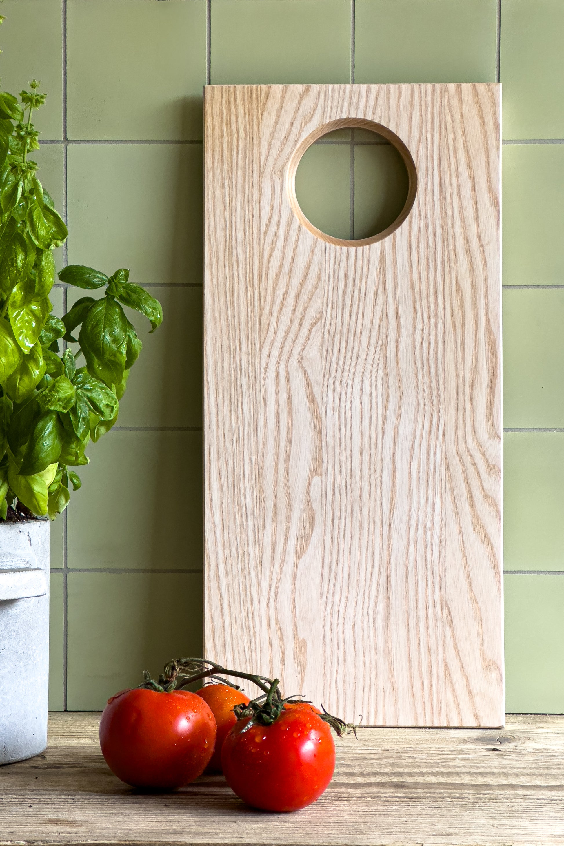 Schneidebrett aus heller Esche mit praktischer Bohrung, perfekt zum Aufhängen in der Küche, vor einer hellgrünen Kachelwand. Daneben steht frischer Basilikum in einem Topf und drei rote Tomaten liegen auf einer Holzoberfläche. Hochwertiges Holz-Schneidebrett für stilvolles Kochen und Zubereiten in einer natürlichen Küchenumgebung. Ein nachhaltiger Küchenhelfer von finbeaux!