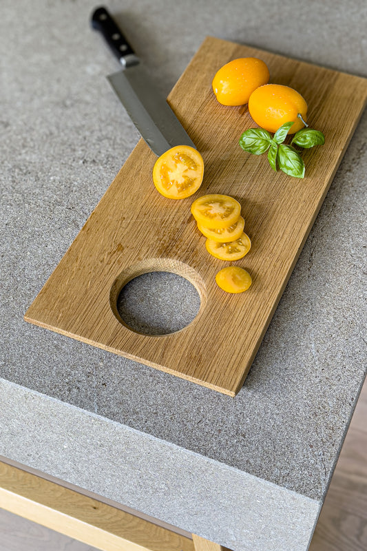 Massives Holz-Schneidebrett auf einer grauen Küchenarbeitsplatte mit geschnittenen gelben Tomaten und Basilikumblättern. Ein scharfes Küchenmesser liegt auf dem Brett, das aus hochwertiger Eiche gefertigt ist und eine praktische Bohrung zum Aufhängen bietet. Perfektes Setup für die Zubereitung frischer und gesunder Zutaten in einer modernen Küche. Du bist es dir Wert!